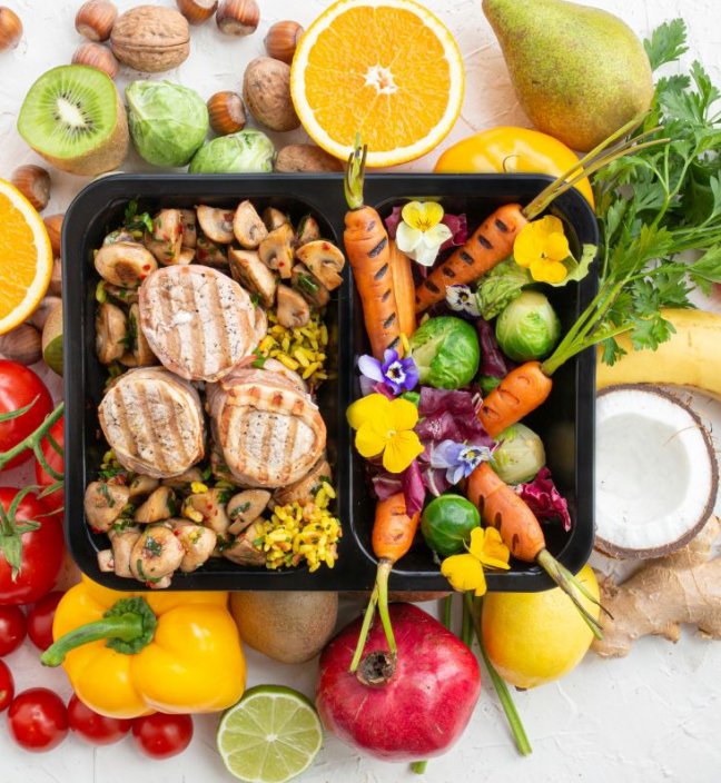 Top view of fruits around a catering plastic box with grilled chicken and vegetables, and saffron rice